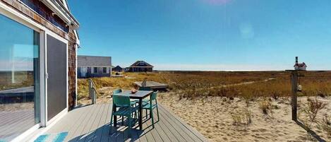 A wooden deck with outdoor furniture, a glass door entrance, and a view of the beach and dunes.