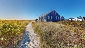 A coastal house with a wooden pathway leading to it, surrounded by tall grasses and a clear sky. - Cozy cottage steps to the beach with patio, W/D, & partial AC (North Truro)