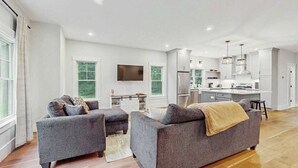 Una sala de estar moderna con sofás grises, un televisor de pantalla plana y una zona de cocina con armarios blancos y una isla central.