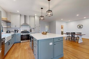 A modern kitchen with a central island, built-in appliances, and a dining area with a wooden table and chairs. - Modern Luxe on the Cape | Sleek Design, Central Air, Serene Views (Mashpee)