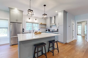 A modern kitchen with a large island, stainless steel appliances, and pendant lighting. - Modern Luxe on the Cape | Sleek Design, Central Air, Serene Views (Mashpee)