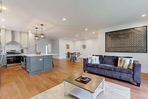 A modern kitchen with a central island, a dining area with a table and chairs, a living room with a sofa and a coffee table, and a large wall-mounted periodic table of elements. - Modern Luxe on the Cape | Sleek Design, Central Air, Serene Views (Mashpee)