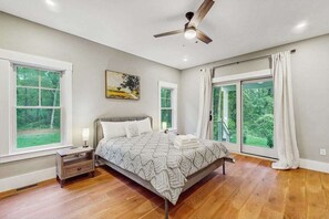 A bedroom with a bed, nightstands, a ceiling fan, large windows, and a view of greenery.
