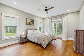 A bedroom with a bed, a nightstand, a ceiling fan, and large windows. - Modern Luxe on the Cape | Sleek Design, Central Air, Serene Views (Mashpee)