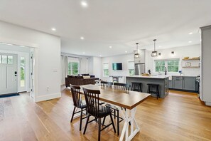 A modern kitchen with a dining area, wooden flooring, and a large window. - Modern Luxe on the Cape | Sleek Design, Central Air, Serene Views (Mashpee)