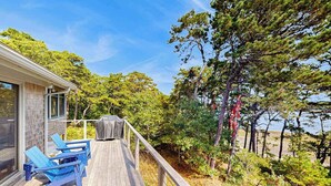 A wooden deck with blue chairs, a covered grill, and a view of trees and a body of water. - Drummer Cove Retreat | House in Wellfleet (Wellfleet)