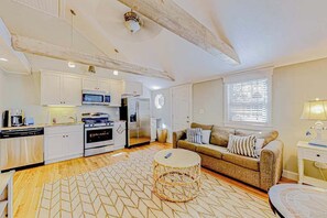 Una cocina moderna con armarios blancos, microondas, estufa y refrigerador. Hay una mesa de centro redonda de mimbre y un sofá color beige con almohadones a rayas.