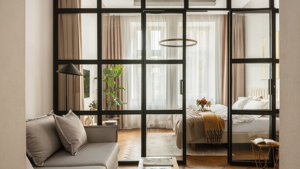 Una sala de estar moderna con sofá, mesa de centro y vistas a un dormitorio con cama y cortinas, separadas por un panel de vidrio.