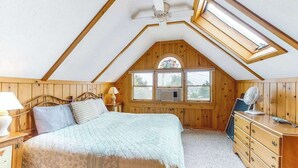 Ein Schlafzimmer mit schrägen Wänden, einem Bett, einem Schminktischchen, einem Nachttisch, einer Lampe, einem Ventilator und einem Fenster mit Blick.