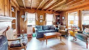 Ein gemütliches Blockhauss-Interieur mit Holzbalkendecke, einer Couch, einem Sessel, einem kleinen Tisch und einem Kamin.