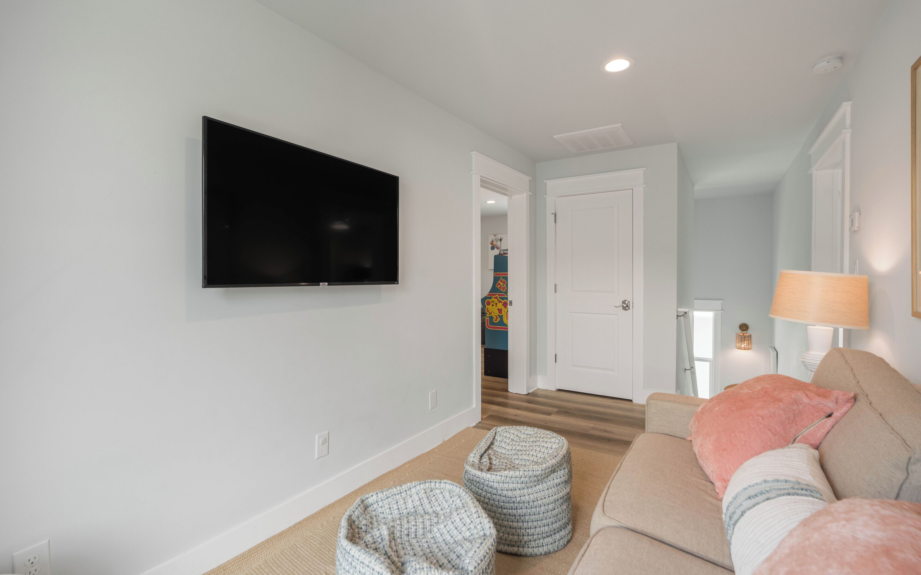 Un salon avec un téléviseur à écran plat fixé au mur, un canapé beige, deux poufs, une lampe et une porte donnant sur une autre pièce.