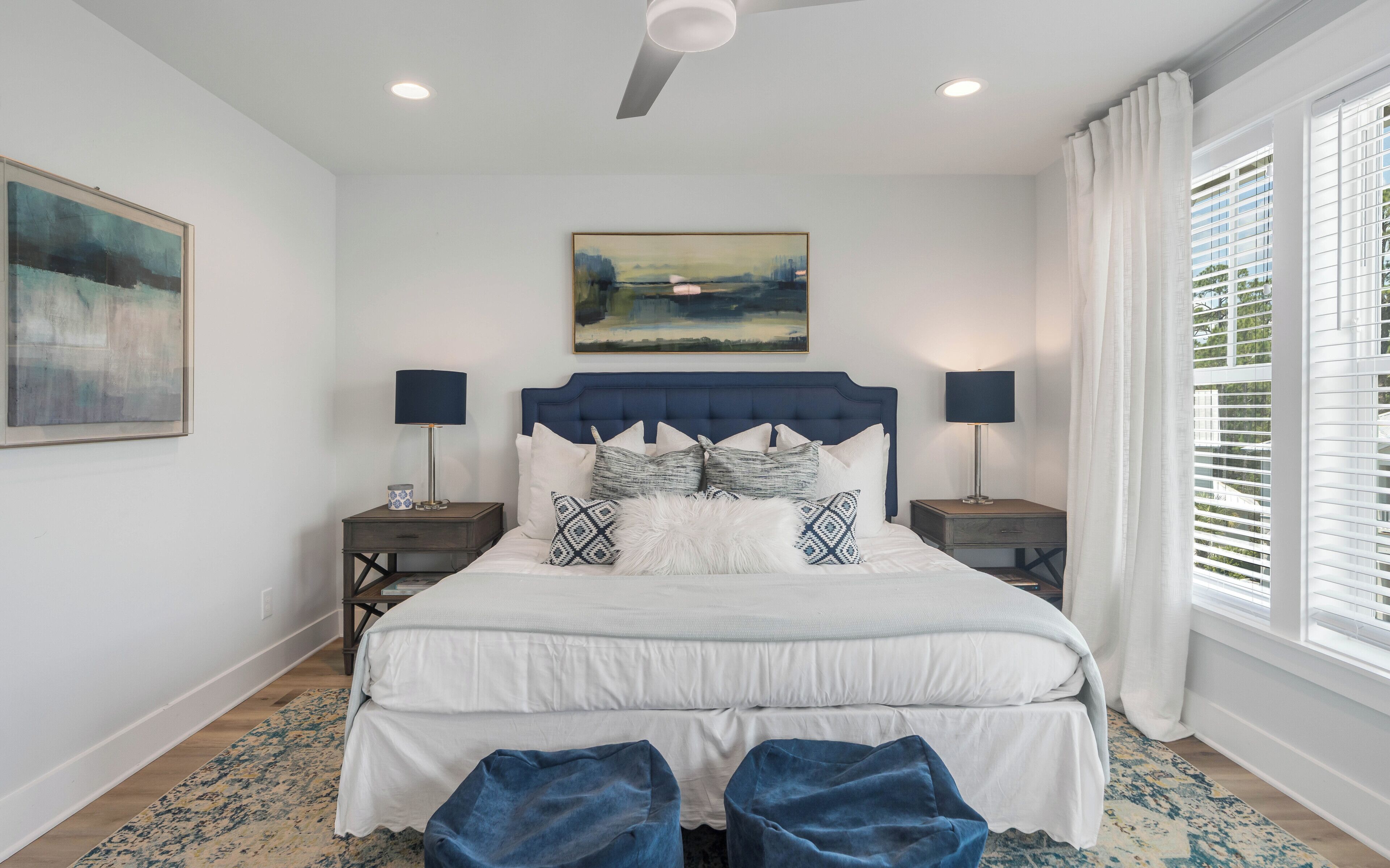 Une chambre à coucher avec un grand lit, deux tables de chevet avec des lampes, un ventilateur de plafond, un tableau au-dessus du lit et une fenêtre avec des stores.