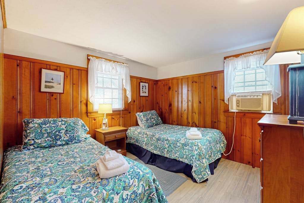 Ein Zimmer mit zwei Betten, Holzwänden, einem Fenster mit Vorhängen, einem Nachttisch mit Lampe und einer Klimaanlage.