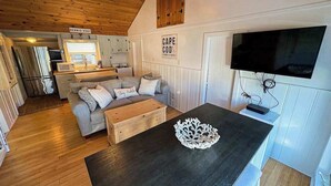 A cozy cabin interior with a kitchenette, a dining area, and a living room with a sofa and a flat-screen TV. - Coastal cottage close to sandy beach with private deck, kayaks & dog friendly (Dennis Port)