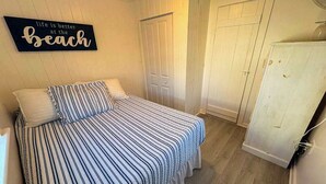A bedroom with a striped bedspread, a wall sign, and a white wardrobe. - Coastal cottage close to sandy beach with private deck, kayaks & dog friendly (Dennis Port)