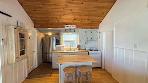 A compact kitchen with a wooden ceiling, a central island with stools, white cabinetry, and a sign reading 'DENNIS PORT'. - Coastal cottage close to sandy beach with private deck, kayaks & dog friendly (Dennis Port)