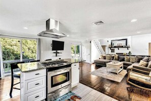A modern kitchen with a stove, oven, and ventilation hood. There is a dining area with a table and chairs, and a living room with a sofa and a coffee table. - Spacious coastal retreat with heated pool, kayaks, game room & pond views (Centerville)