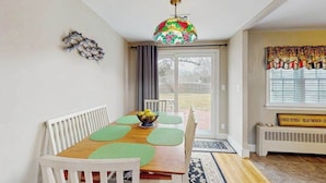 En matplats med ett träbord, gröna bordsunderlägg, vita stolar, en färgstark hängande lampa och utsikt genom en glasdörr.