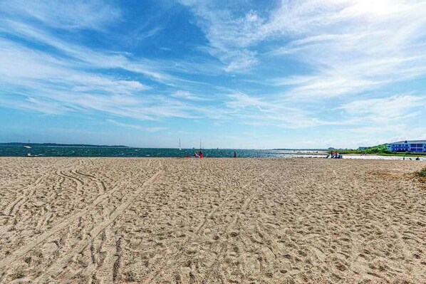 A sandy beach with visible tire tracks, a clear blue sky, and distant boats on the water. - Private house close to beach with grill, AC, yard, and deck (Yarmouth)