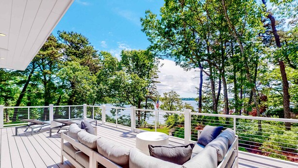 A deck with outdoor seating, a round table, and a view of trees and water. - Luxe lakefront home on Follins Pond with private beach, amazing decks & yard (Yarmouth Port)