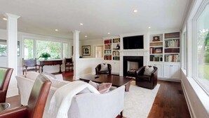 A spacious living room with a fireplace, bookshelves, and a television. - Luxe lakefront home on Follins Pond with private beach, amazing decks & yard (Yarmouth Port)