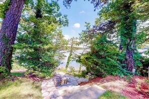 A stone pathway leading through a forested area with a lake in the background. - Luxe lakefront home on Follins Pond with private beach, amazing decks & yard (Yarmouth Port)