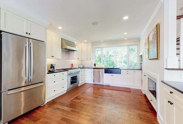 A modern kitchen with white cabinetry, stainless steel appliances, and a large window providing natural light. - Luxe lakefront home on Follins Pond with private beach, amazing decks & yard (Yarmouth Port)