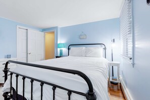 A bedroom with a bed, a nightstand, a lamp, and a window with blinds. - Cape Charmer | Walk to Market, Golf & Eateries | New Kitchen & Big Backyard (Yarmouth Port)