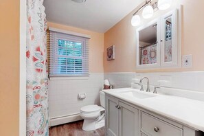 A bathroom with a toilet, sink, mirror, and window with blinds. - Cape Charmer | Walk to Market, Golf & Eateries | New Kitchen & Big Backyard (Yarmouth Port)