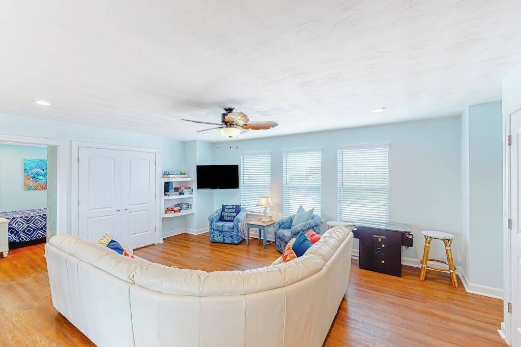 A living room with a white sectional sofa, a ceiling fan, a flat-screen TV, and a window with blinds.
