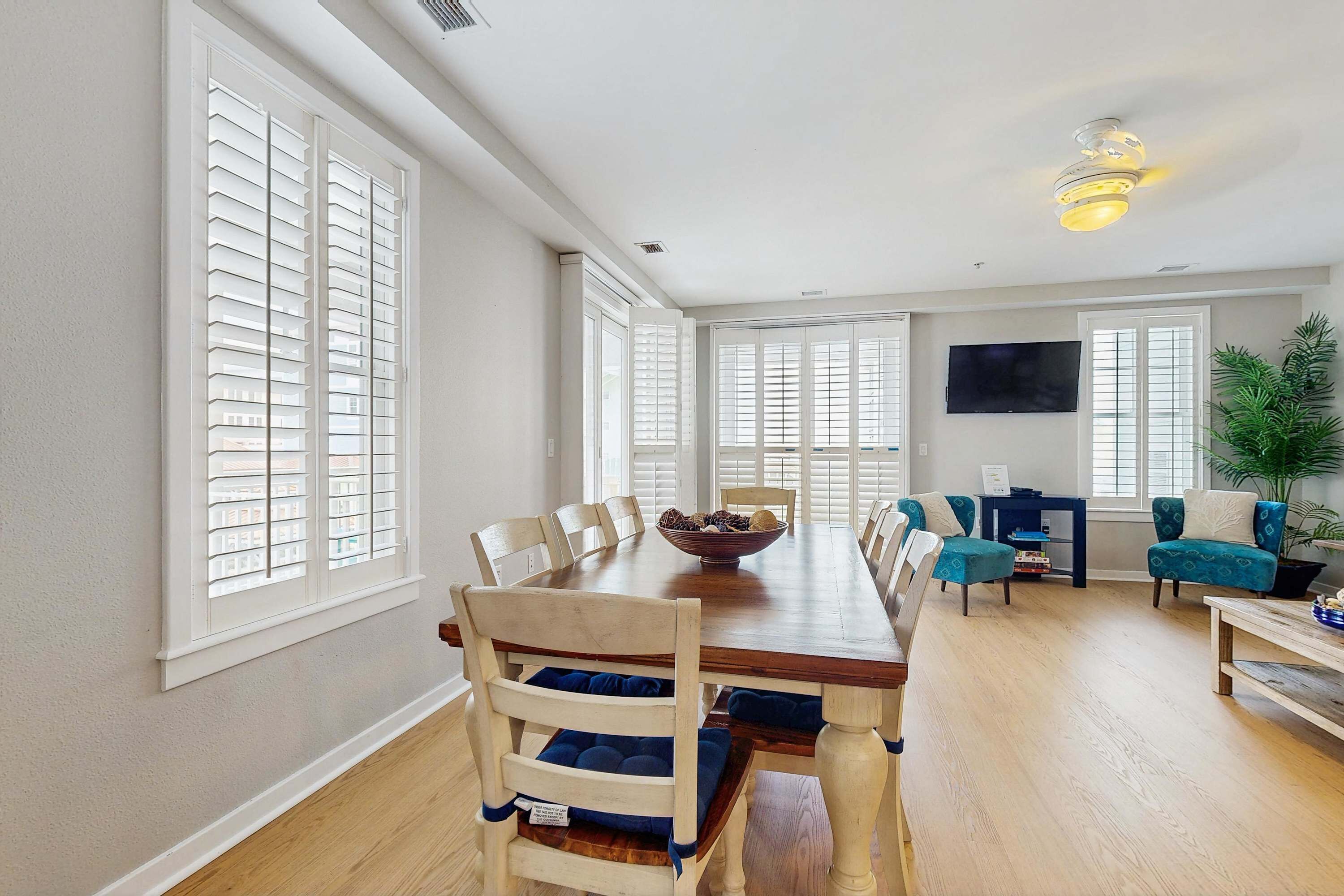 A spacious dining area with a wooden table and chairs, a large window with blinds, a television mounted on the wall, and a potted plant.