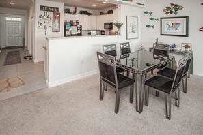 Un coin repas avec une table à plateau en verre et quatre chaises, une cuisine avec des meubles blancs et un mur décoré sur le thème des poissons.