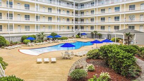 Ein mehrstöckiges Hotel mit einem zentral gelegenen Swimmingpool, Sitzmöglichkeiten im Freien und einem angelegten Garten.