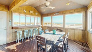 Eine Holzdachterrasse mit Tisch, Stühlen, einem Deckenventilator und großen Fenstern mit Blick auf den Strand.