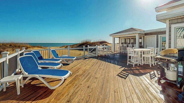 Eine Holzterrasse mit Liegestühlen und Grillplatz, mit Blick auf einen Strand.