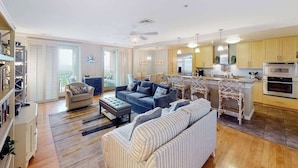 Een ruime woonkamer met een bank, fauteuil en poef, een keuken met moderne apparatuur, en een eethoek met een bar.