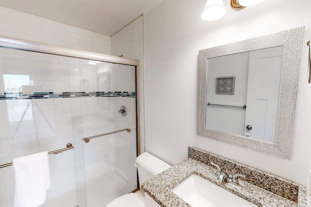 A bathroom with a glass shower enclosure, a white toilet, a granite countertop sink, and a mirror with a decorative frame.