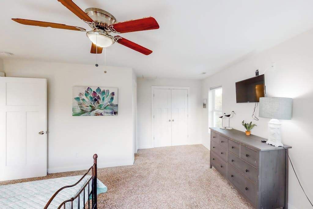 Une chambre à coucher avec un ventilateur de plafond, un lit, une commode, un tableau et une porte.