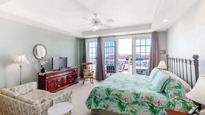 A hotel room with a bed, a TV, a sofa, a chair, a balcony with a view, and a ceiling fan. - Second-floor, ocean view condo with balcony, foosball, WiFi & on-site pool/gym (Virginia Beach)