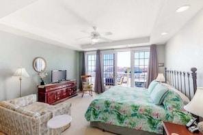 A hotel room with a bed, a TV, a sofa, a chair, a balcony with a view, and a ceiling fan. - Second-floor, ocean view condo with balcony, foosball, WiFi & on-site pool/gym (Virginia Beach)
