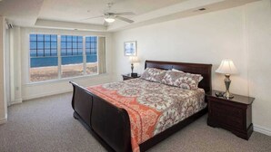 A bedroom with a bed, two bedside tables, a ceiling fan, a window with a beach view, and a framed picture on the wall. - Oceanfront, corner condo with wrap-around deck, great views & on-site pool/gym (Virginia Beach)