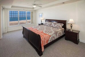 A bedroom with a bed, two bedside tables, a ceiling fan, a window with a beach view, and a framed picture on the wall. - Oceanfront, corner condo with wrap-around deck, great views & on-site pool/gym (Virginia Beach)