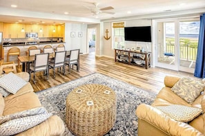 A spacious living area with a dining set, a large round woven ottoman, and a balcony with a view. - Bay-view condo w/Free Wifi,  pools, outdoor shower, gym, lanai & beach access (Virginia Beach)