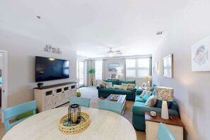 A living room with a TV, teal sofa, and a dining area with a round table. - Sunny beach condo with washer/dryer, pool/hot tub & gym (Virginia Beach)