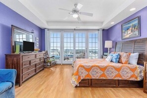 A bedroom with a bed, dresser, TV, and a view through large windows. - Ocean View Condo in an Oceanfront Property with Balcony, WiFi & Shared Pool/Gym (Virginia Beach)