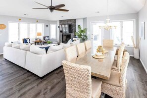 A modern living room with a sofa, dining table, and chairs, a fireplace, and large windows. - Beautiful oceanside home with pool, beautiful decor, WiFi, washer/dryer, AC (Virginia Beach)