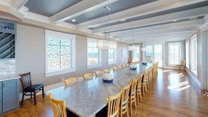 A spacious dining area with a long table, multiple chairs, pendant lights, and large windows. - Wonderful beachfront home w/private indoor & outdoor pools, WiFi, AC, deck (Virginia Beach)