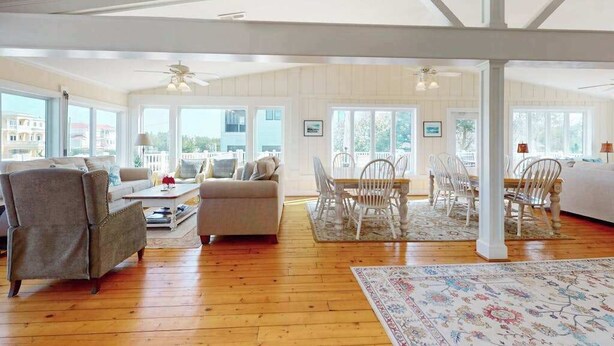 A spacious living room with a high ceiling, wooden flooring, a dining area with white chairs, and large windows offering views. - 8 BDR & 7.5 BA Luxury Beach House w/private pool & hot tub (Virginia Beach)