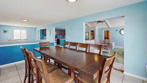 Un grand espace à manger avec une longue table en bois et des chaises, une cuisine à l’arrière-plan et un coin salon avec un canapé et un ventilateur de plafond.