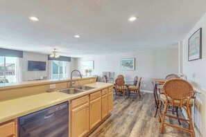 A kitchen with wooden cabinets, a dishwasher, a dining area with wicker chairs, and a ceiling fan. - Sunny Condo w/Shared Outdoor Pool, Central A/C, Private W/D - Near the Ocean (Virginia Beach)